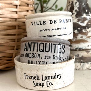 May include: A stack of three round ceramic crocks with black printed text. The top crock reads VILLE DE PARIS NOUVEAUTES, the middle reads ANTIQUITES Juste COLSON, rue Gretry, 40, BRUXELLES, and the bottom crock reads French Laundry Soap Co.