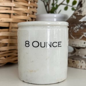May include: A small, off-white ceramic jar with the text "8 OUNCE" in black. The jar is cylindrical with a slightly rounded top and is likely a vintage kitchen item. The background includes a wicker basket and decorative items.