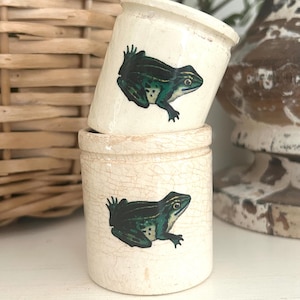 May include: Two off-white ceramic jars with a frog illustration. The frogs are green and black. The jars have a distressed, crackled finish. One jar is stacked on top of the other.