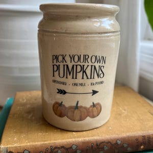 May include: A beige ceramic jar with the text "PICK YOUR OWN PUMPKINS" and illustrations of pumpkins. The jar is sitting on top of a stack of old books. The text also includes "OPEN DAILY. ONE MILE. 10¢ POUND."