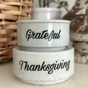 May include: A two-tiered ceramic dish with a white crackle glaze finish. The top tier features the word Grateful in black script, while the bottom tier displays the word Thanksgiving in black script. The object has a cylindrical shape.