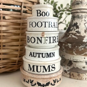 May include: A stack of six off-white ceramic bowls with a crackled finish. The bowls feature black text reading Boo, FOOTBALL, BONFIRE, AUTUMN, and MUMS, while the base bowl displays a repeating pattern of black bats around the exterior.