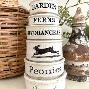 May include: A stack of six white ceramic ramekins with black text and illustrations. The labels read GARDEN, FERNS, HYDRANGEAS, Peonies, and Boxwood, with a central ramekin featuring a black hare illustration. Each piece has a smooth finish.