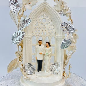 May include: A vintage wedding cake topper featuring a bride and groom figurine standing in front of a white arched structure with a decorative window. The topper is adorned with silver leaves and white floral accents, and two silver bells.