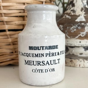 May include: A white ceramic mustard jar with black text that reads "MOUTARDE P. JACQUEMIN PERE & FILS MEURSAULT COTE D'OR". The jar has a rounded body and a narrow neck. The background includes a wicker basket and a decorative vase.