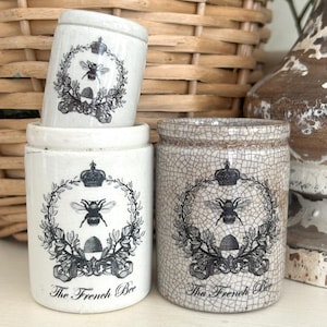 May include: A collection of ceramic jars with bee-themed designs. The jars are white and cream-colored, with black and gray illustrations of bees, crowns, and floral wreaths. The text "The French Bee" is printed on the front of the jars.