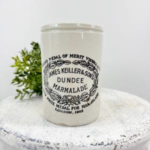 May include: Cream-colored antique marmalade jar with black text and a decorative border. The jar reads "James Keiller & Son's Dundee Marmalade" and "Grand Medal of Merit Vienna 1873". A vintage kitchenware item.