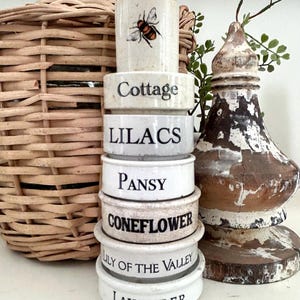 May include: A stack of white ceramic containers with black lettering, including "Cottage," "Lilacs," "Pansy," "Coneflower," "Lily of the Valley," and "Lavender." The top container features a bee illustration. A woven basket and a decorative wooden finial are also present.