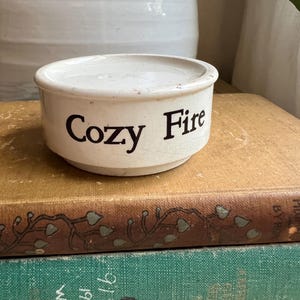 May include: A small, off-white ceramic bowl with the words "Cozy Fire" in black lettering. The bowl is sitting on top of two antique books with brown and green covers. The background includes a white ceramic vase.