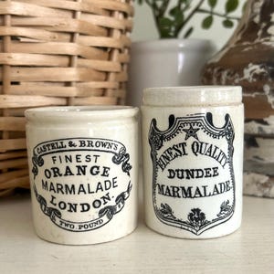 May include: Two off-white ceramic marmalade jars with black printed labels. The left jar reads Castell & Brown's Finest Orange Marmalade London Two Pound. The right jar reads Finest Quality Dundee Marmalade with a star and crest design.