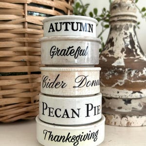 May include: A stack of five white ceramic ramekins with black text. The labels read AUTUMN, Grateful, Cider Donut, PECAN PIE, and Thanksgiving. Each ramekin has a smooth, glazed finish, with the middle one showing a subtle crackle texture.