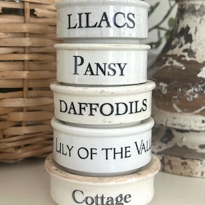 May include: A stack of five small, off-white ceramic containers with black lettering. The containers are labeled "Lilacs," "Pansy," "Daffodils," "Lily of the Val," and "Cottage." The containers have a rustic, vintage aesthetic.