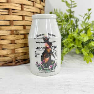 May include: A white ceramic jar with a black and brown illustration of a dog wearing a hat, surrounded by pink flowers. The jar has text in black, including the word "Paris". The jar is next to a woven basket and green foliage.