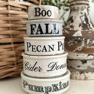 May include: A stack of five off-white ceramic ramekins with a crackled glaze finish. Each ramekin features black text: Boo, FALL, PECAN PIE, Cider Donuts, and Pumpkins. The stack is arranged with the smallest on top and the largest on the bottom.