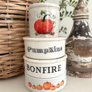 May include: A stack of four cylindrical ceramic containers with a cream-colored finish. The top container features a painted orange pumpkin. The middle containers display the black text Pumpkins and BONFIRE. The bottom container has small pumpkins.