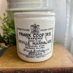 May include: A vintage white ceramic jar with black text reading "Frank Cooper's Oxford Seville Marmalade." The jar has a cylindrical shape with a small lip and is sitting on a book. The text includes the words "Home Made" and "Registered Trade Mark."