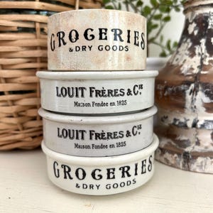 May include: A stack of four small, round ceramic containers. The top and bottom containers are off-white with the words "GROCERIES & DRY GOODS" in black. The middle two containers are white with "LOUIT FRÈRES & C" and "Maison Fondée en 1825" in black.