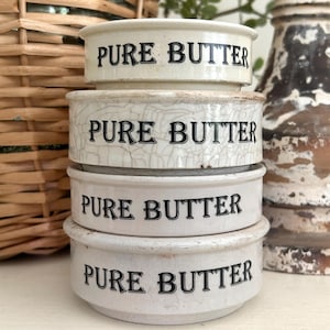 May include: A stack of four round ceramic butter crocks, each featuring the text PURE BUTTER in black lettering. The crocks have a cream-colored glaze, with the second one from the top displaying a distinct brown crackle finish across its surface.