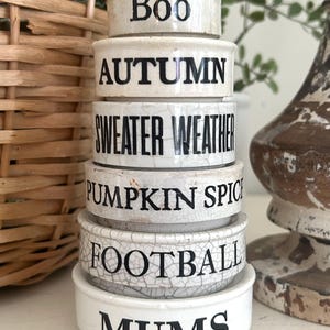 May include: A stack of six white ceramic bowls with a crackled glaze finish. Each bowl features black text: Boo, Autumn, Sweater Weather, Pumpkin Spice, Football, and Mums. The bowls are arranged in a vertical column from smallest to largest.