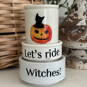 May include: A stack of three white ceramic containers. The top piece features a black cat inside an orange jack-o'-lantern. The middle piece has the black text Let's ride, and the bottom piece has the black text Witches! printed on the front.