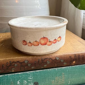 May include: A small, off-white ceramic bowl with a lid, decorated with a row of orange pumpkins. The bowl has a cracked, aged appearance. The bowl is sitting on top of a stack of vintage books.