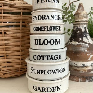 May include: A stack of seven white ceramic bowls with black text printed on each. The text reads FERNS, HYDRANGEAS, CONEFLOWER, BLOOM, COTTAGE, Sunflowers, and GARDEN from top to bottom. Each bowl has a smooth, rounded rim and a glossy finish.