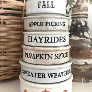 May include: A stack of six round, off-white ceramic bowls with a crackled finish. Each bowl features black text: FALL, APPLE PICKING, HAYRIDES, PUMPKIN SPICE, and SWEATER WEATHER. The bottom bowl is decorated with a row of small, orange pumpkin illustrations.