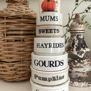 May include: A stack of six white ceramic rings with black text. The top piece features a painted orange pumpkin. The rings are labeled with the words MUMS, SWEETS, HAYRIDES, GOURDS, Pumpkins, and CORNSTALKS in varying black fonts and styles.