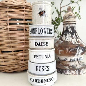 May include: A stack of small, white ceramic pots with black text labels: SUNFLOWERS, DAISY, PETUNIA, ROSES, and GARDENING. The top pot has a bee illustration. A decorative, distressed wooden object and a wicker-covered bottle are in the background.