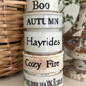 May include: A stack of five off-white ceramic ramekins with a crackled glaze finish. Each ramekin features black printed text: Boo, AUTUMN, Hayrides, Cozy Fire, and Pumpkins. The cylindrical containers are stacked vertically in a single column.