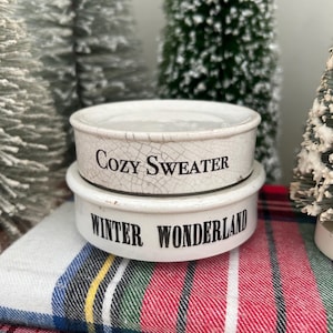 May include: Two white ceramic bowls with black text, one reading "Cozy Sweater" and the other "Winter Wonderland." The bowls are stacked on a red, green, and blue plaid fabric, with miniature Christmas trees in the background.