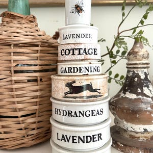 May include: A stack of seven white ceramic measuring cups with black text and illustrations. The cups feature the words Lavender, Cottage, Gardening, Hydrangeas, Lavender, and Peonies, along with a bee illustration and a running rabbit graphic.