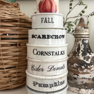 May include: A stack of six white ceramic containers with a crackled finish. The top piece features a pumpkin illustration. The others are labeled with black text: FALL, SCARECROW, CORNSTALKS, Cider Donuts, and Pumpkins, with small pumpkins at the base.