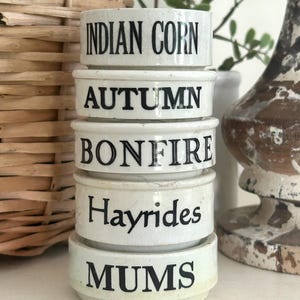 May include: A stack of five cylindrical white ceramic containers with black text. The labels read INDIAN CORN, AUTUMN, BONFIRE, Hayrides, and MUMS from top to bottom. Each container has a smooth finish and a slightly raised rim at the base.