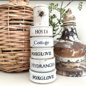 May include: A stack of white ceramic plant markers with black text, including "HOSTA", "COTTAGE", "FOXGLOVE", and "HYDRANGEA". The top marker features a bee illustration. A woven bottle and decorative wooden piece are in the background.