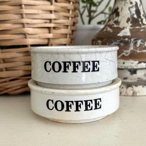 May include: Two stacked, round, off-white ceramic bowls with a visible crackle glaze finish. Each bowl features the word COFFEE printed in bold, black, sans-serif capital letters on the front. The bowls have a simple, low-profile design.