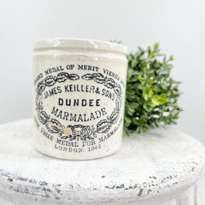 May include: A vintage Dundee Marmalade jar with a cream-colored base and black lettering. The jar features the text "JAMES KEILLER & SONS DUNDEE MARMALADE" within a decorative wreath. Additional text includes "GRAND MEDAL OF MERIT VIENNA" and "PRIZE MEDAL FOR LONDON. 1862".
