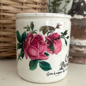 May include: A cylindrical white ceramic pot featuring a floral design of two large pink roses with green leaves and stems. The side of the pot displays black printed text that reads "Chocolat Poulain" and "Goutez & comparez". The surface shows fine cracks.