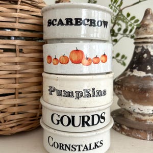 May include: A stack of five off-white ceramic ramekins with a speckled finish. The bowls feature black printed text reading SCARECROW, Pumpkins, GOURDS, and CORNSTALKS, with one bowl displaying a row of orange pumpkin illustrations in the center.