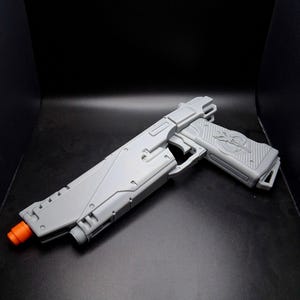 Puede incluir: Una pistola de juguete gris con punta naranja, sobre un fondo negro. La pistola presenta un diseño detallado con empuñadura texturizada y varios componentes mecánicos. La estética general sugiere un tema futurista o de ciencia ficción.