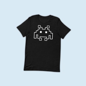 May include: A black t-shirt with a white pixelated alien graphic. The alien has a simple design with two eyes and a mouth.