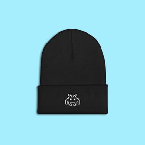 Op de afbeelding: Zwarte beanie met een wit gepixeld alien-ontwerp. De hoed is gemaakt van een zacht, gebreid materiaal en heeft een omgeslagen manchet. De beanie staat tegen een lichtblauwe achtergrond.