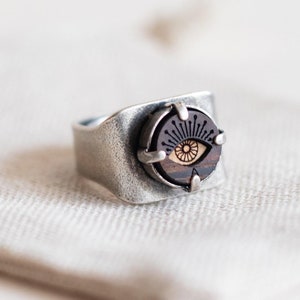 Könnte beinhalten: Ein silberner Ring mit einer dunklen Holzeinlage, die ein stilisiertes Auge zeigt. Das Auge hat eine hellbraune Iris und eine schwarze Pupille.