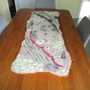 May include: A white felted table runner with abstract designs in pink, green, and gray. The runner has a textured surface and is decorated with small white circles and triangles.