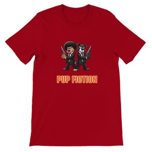 May include: Red t-shirt featuring a cartoon illustration of two dogs in black suits, holding guns. The text "PUP FICTION" is displayed in a yellow and orange font below the dogs. The shirt is made of a soft material.