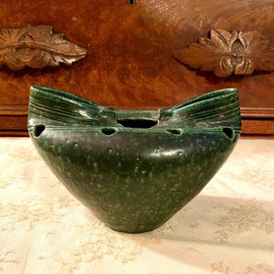 May include: A dark green, speckled, ceramic bowl with a unique shape. The bowl has a wide, curved top with three small holes and a central opening. The bowl's sides taper to a point, and it sits on a patterned surface.