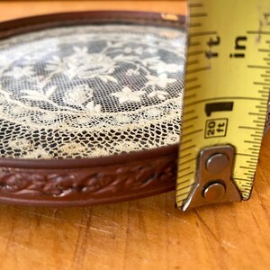 May include: A small, round, brown tray with a glass top. The tray has a decorative, floral design around the edge. The glass top features a delicate, white lace design. A measuring tape is used to show the size of the tray, which is approximately 6 inches in diameter.