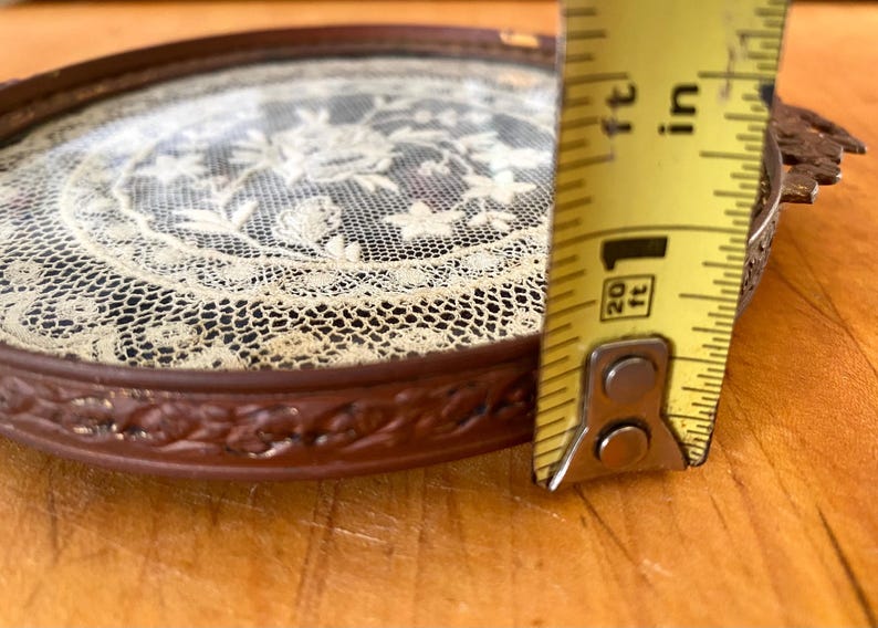 May include: A small, round, brown tray with a glass top. The tray has a decorative, floral design around the edge. The glass top features a delicate, white lace design. A measuring tape is used to show the size of the tray, which is approximately 6 inches in diameter.