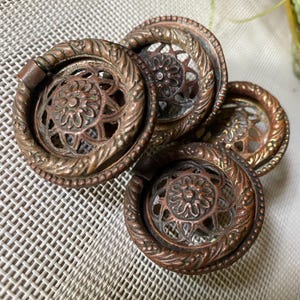 May include: A set of three circular bronze-toned metal drawer pulls featuring a central floral motif surrounded by an openwork lattice pattern. Each pull has a thick, twisted rope-style outer ring and a hinged drop handle for easy gripping.