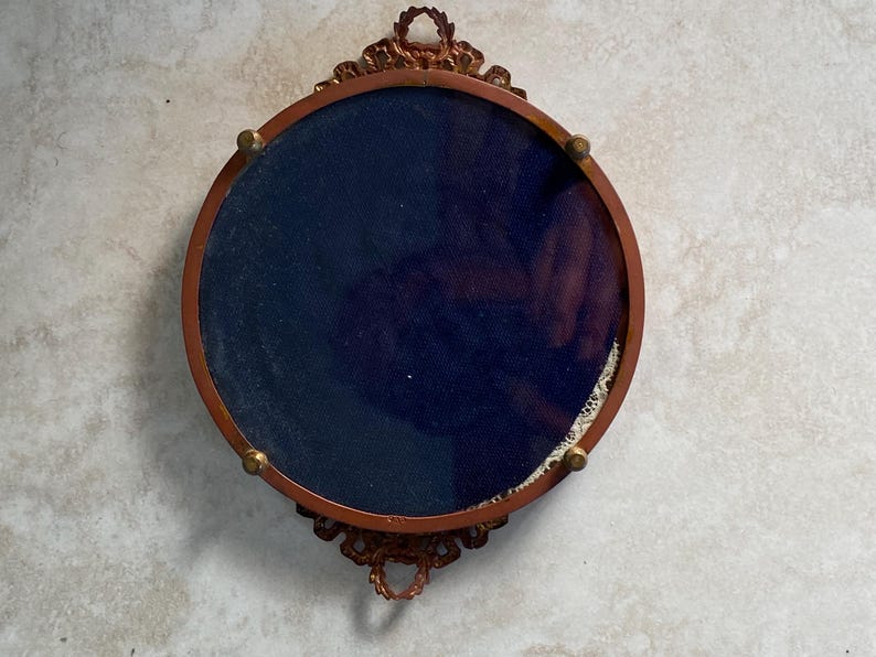 May include: A round, ornate, gold-colored frame with a dark blue velvet backing. The frame has a decorative design with intricate details.